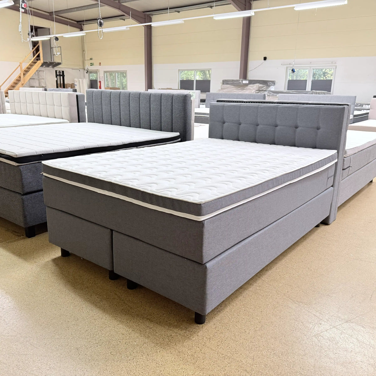 Boxspringbett Jona 140x200 in Steingrau mit gestepptem Kopfteil, Knopfheftung, Kaltschaumtopper und runden schwarzen Holzfüßen, schräge Vorderansicht
